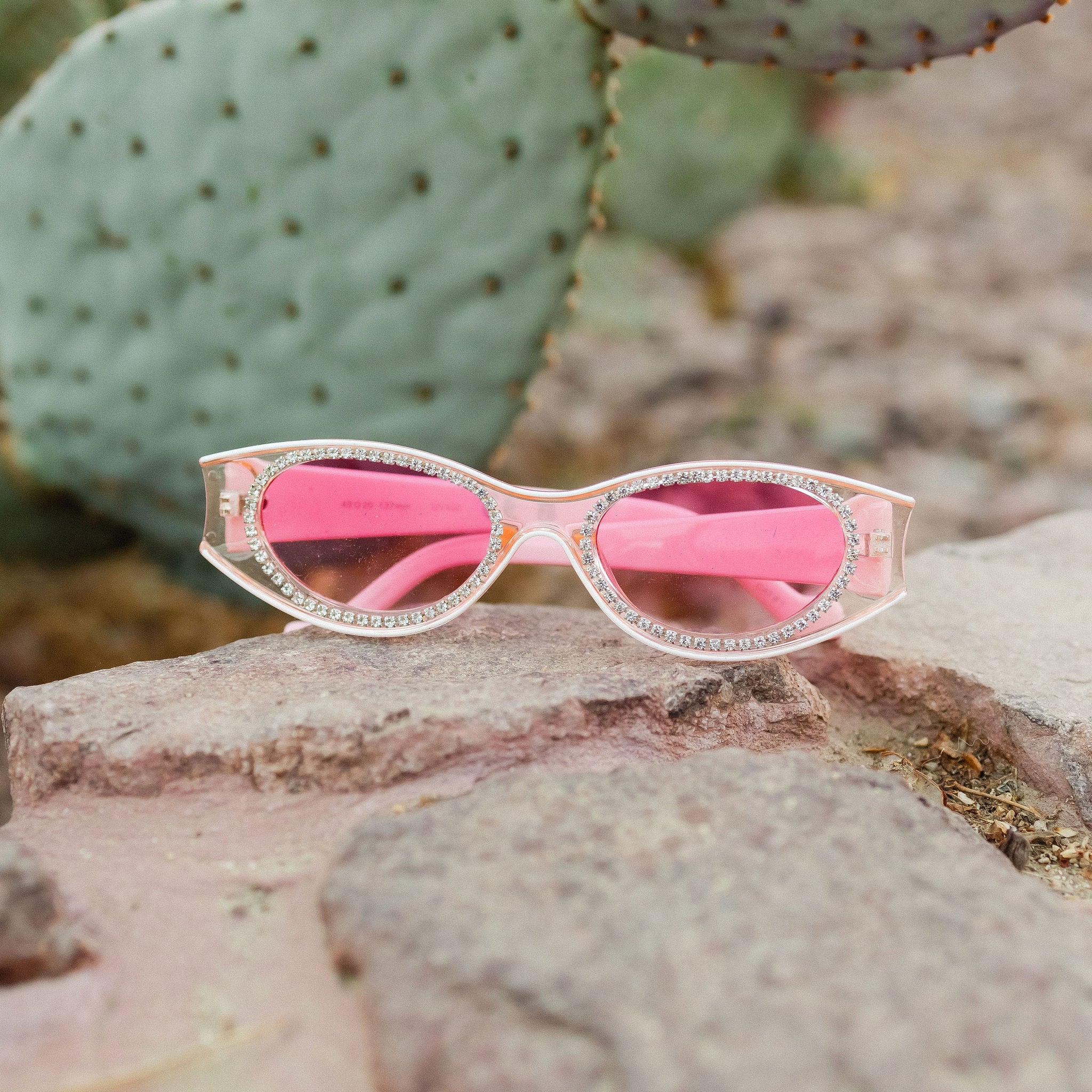 A pair of pink-frame sunglasses with clear lenses and rhinestone embellishments on the lenses.