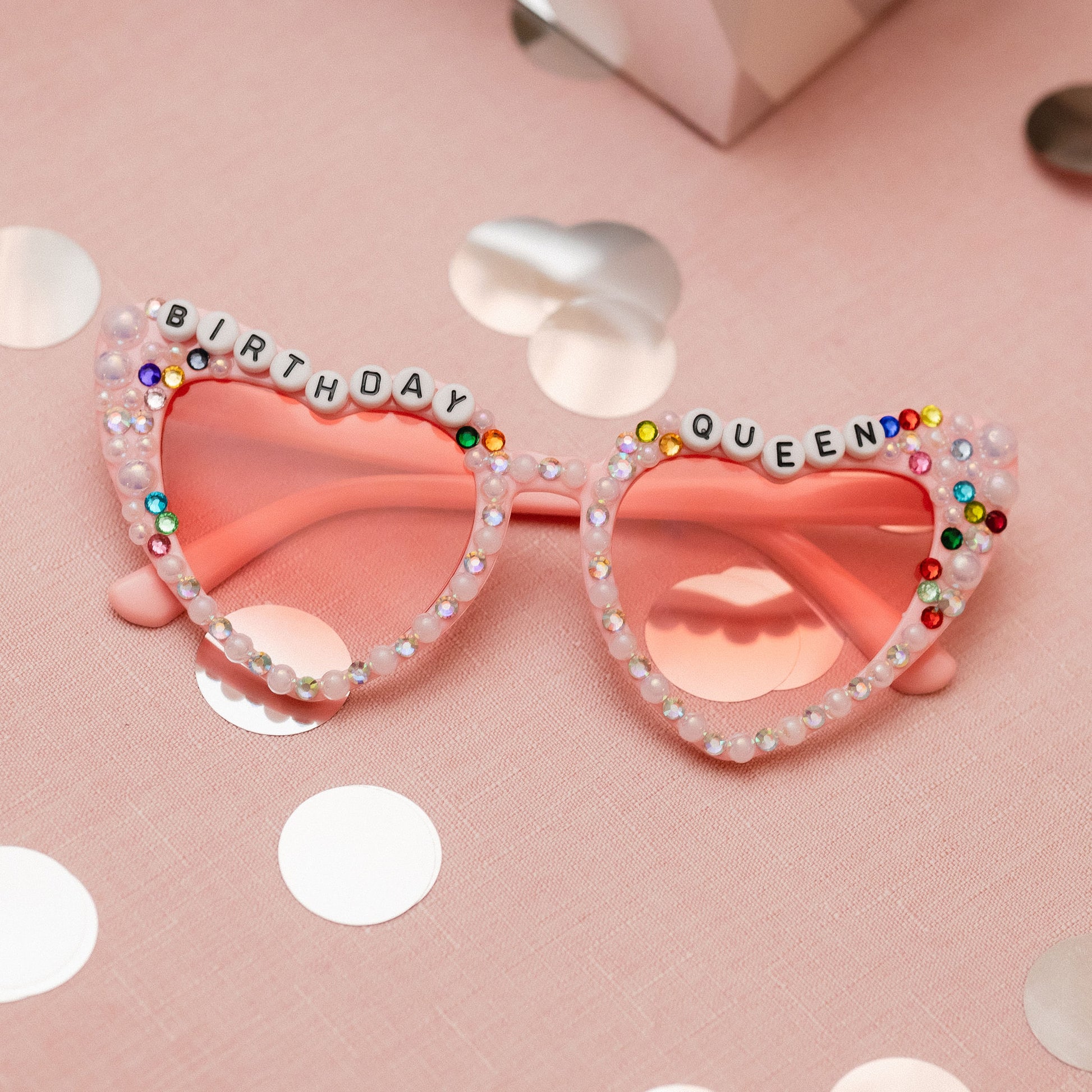 A pair of pink heart-shaped sunglasses with 'BIRTHDAY QUEEN'  multicolored crystals.